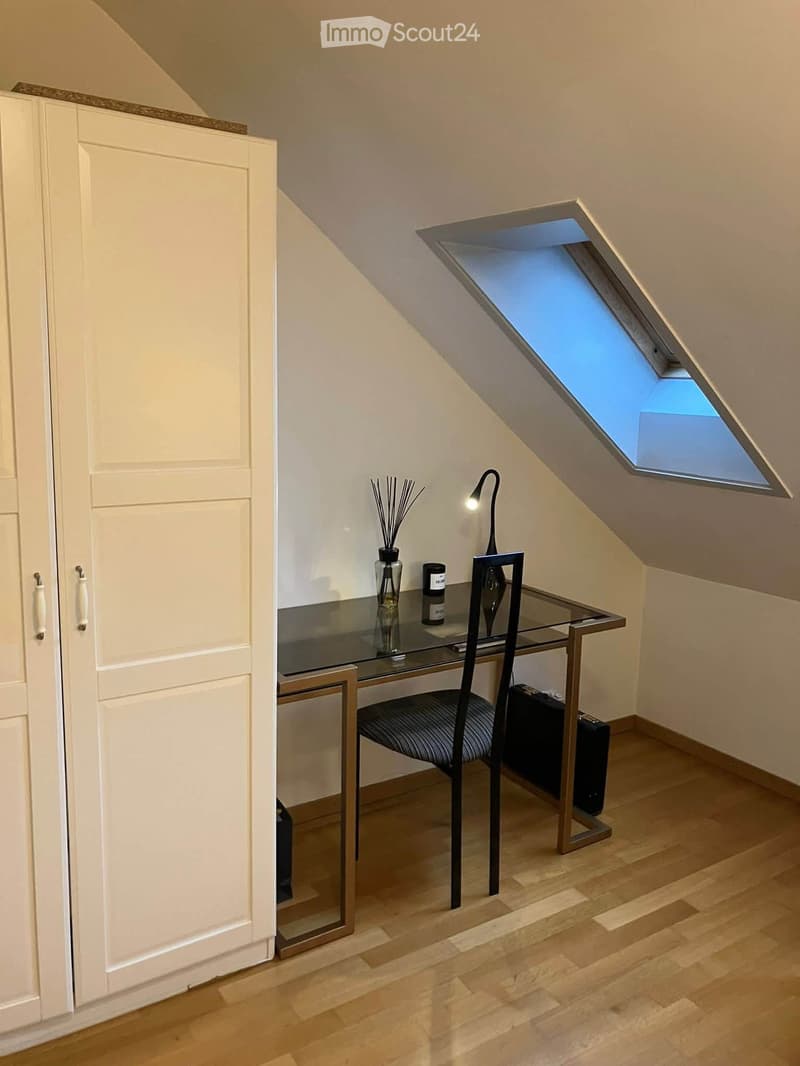 A small, well-lit room with wooden flooring, a glass desk, a chair, a vase with decorative sticks, and a modern lamp. The room features a skylight window and built-in white cabinetry.