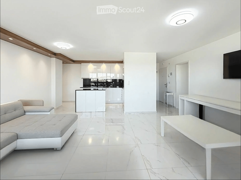 Modern living room with white walls, white marble flooring, a grey sectional sofa, a white coffee table, a kitchen island, and a television mounted on the wall.