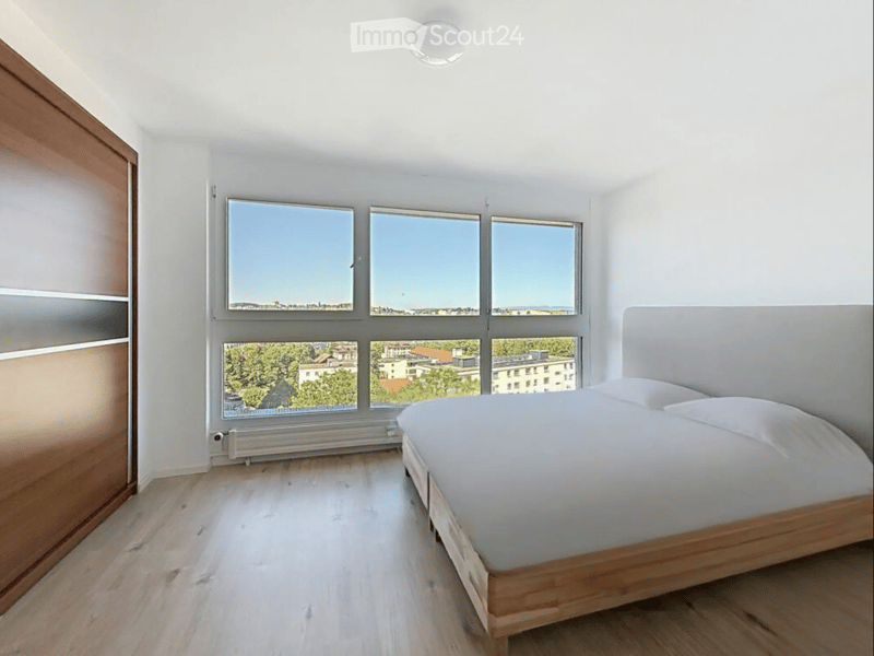 Bright bedroom with wooden flooring, large windows offering a scenic view, a neatly made bed with a headboard, and a modern radiator below the windows.
