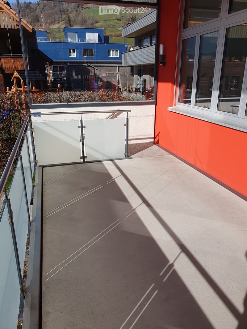 Balcony with glass railing, concrete floor, and view of surrounding houses and greenery.
