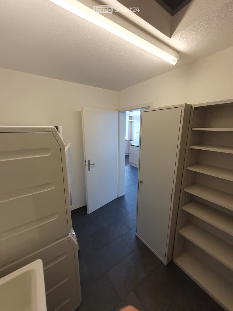 Empty room with tiled floor, white walls, and built-in shelving units. An open door leads to another room with a visible washing machine.