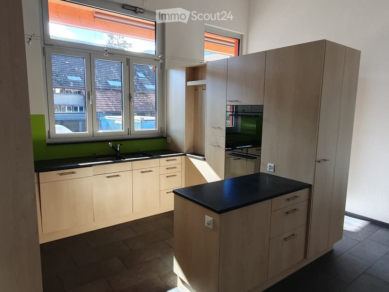 Modern kitchen with light wood cabinets, black countertops, and stainless steel appliances. Features a large window with a view of the roof and surrounding buildings.