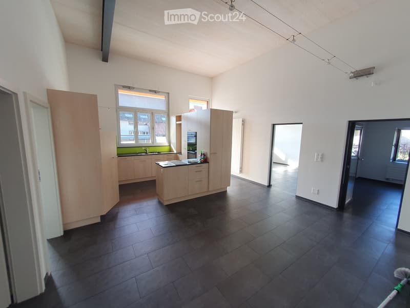 Modern kitchen with light wooden cabinets, dark tiled floor, and stainless steel appliances. Features a central island with sink and faucet, and a window overlooking the outside.
