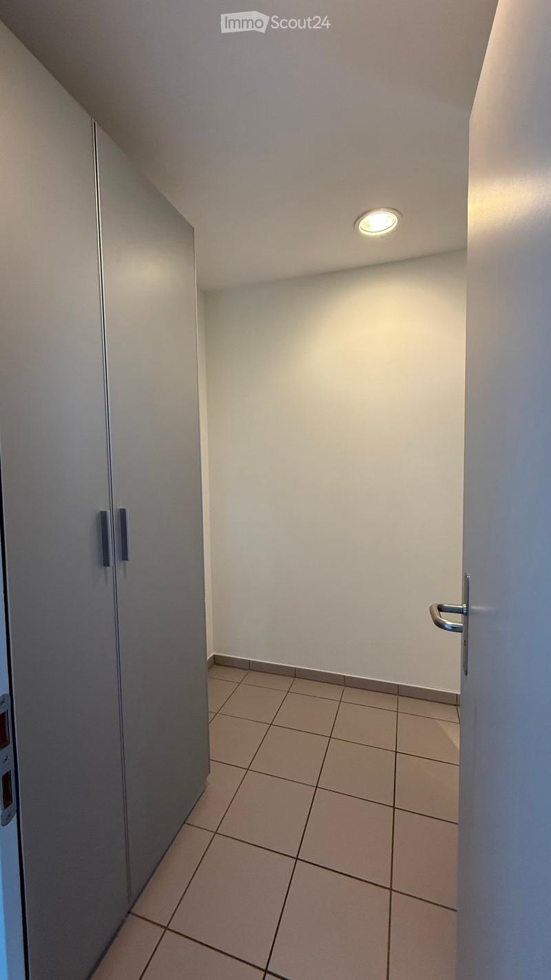 A narrow hallway with tiled flooring, white walls, and a single recessed ceiling light. A closed door is visible on the right, and a cabinet with handles is on the left.