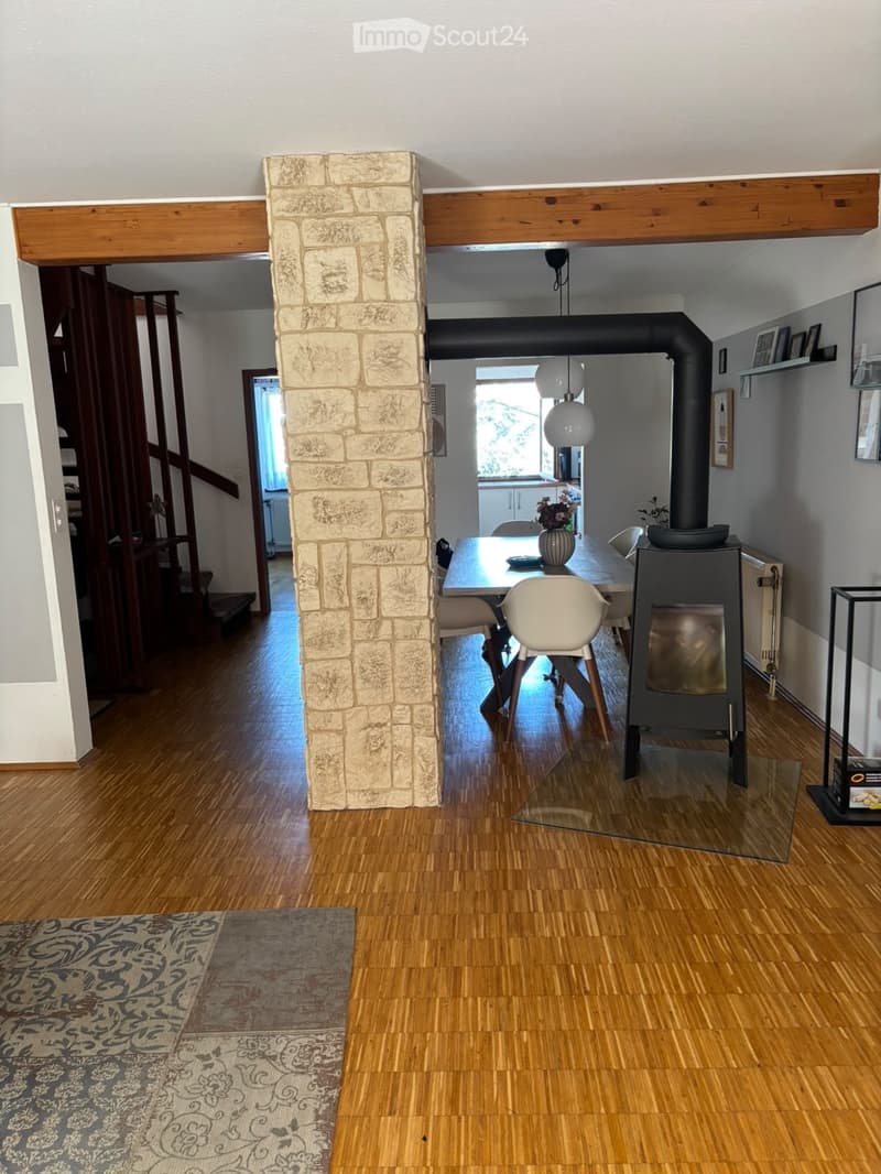 Open-plan living and dining area with wooden flooring, a central stone pillar, a dining table with chairs, a wood stove, and a staircase leading to another level.