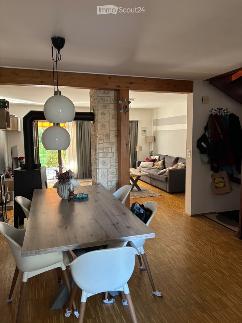 Open-plan living and dining area with wooden flooring, a large dining table with chairs, a sofa with cushions, a coffee table, and decorative items including a vase and candle.