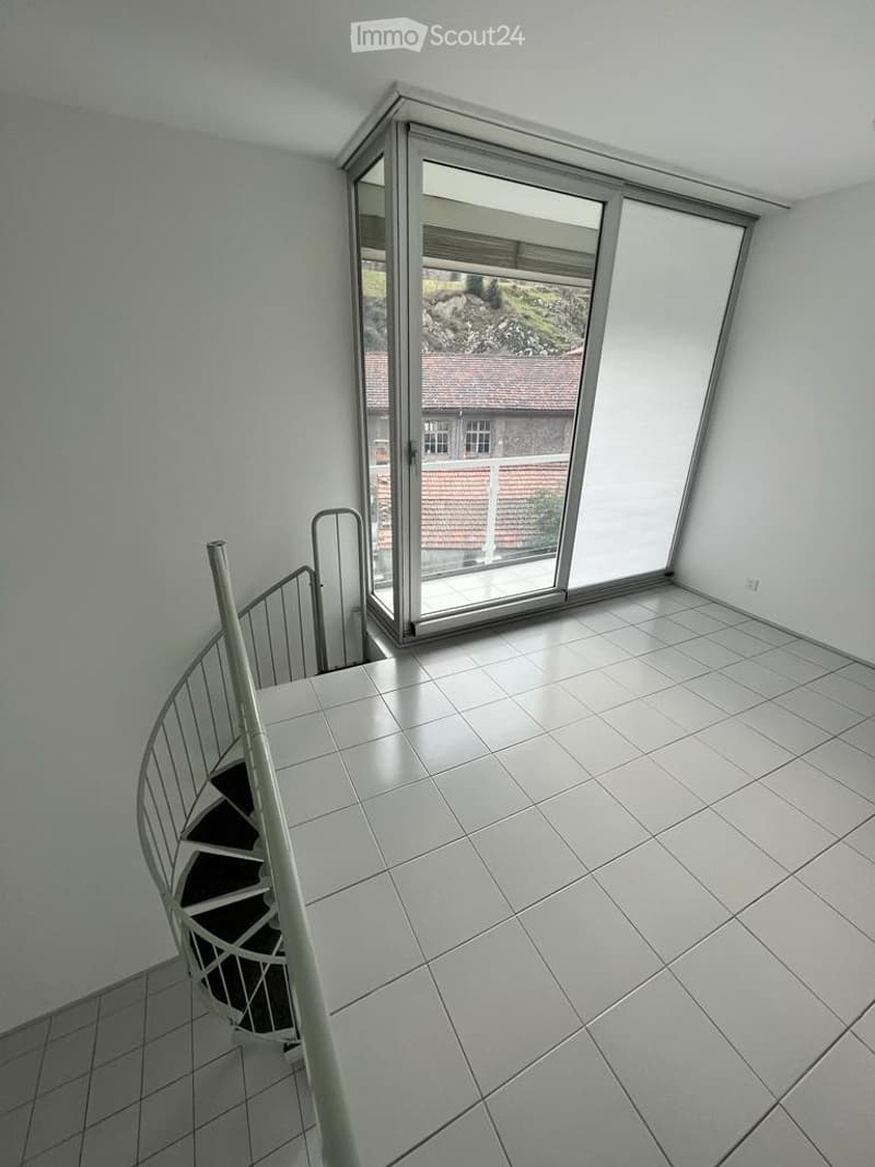 Empty room with white tiled flooring, a modern spiral staircase, and large glass sliding doors leading to a view of a house and greenery outside.