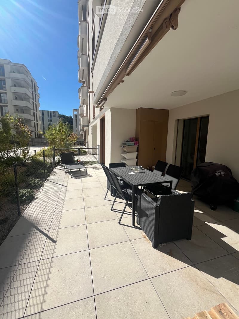 Balcony with tiled floor, outdoor furniture including a table, chairs, and an umbrella, with a view of neighboring buildings and greenery.