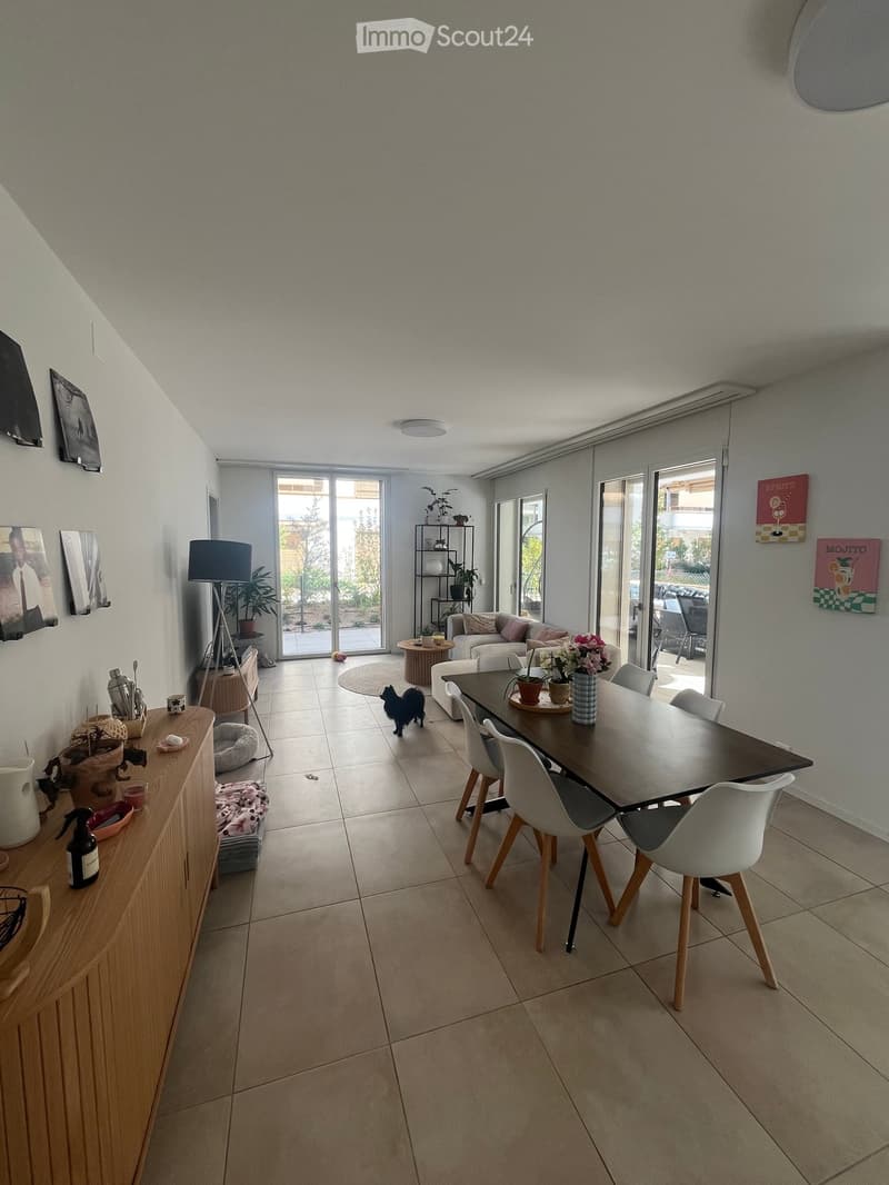 Modern living and dining area with a sofa, dining table, chairs, lamp, cabinet, vase, and picture frames. The space features large windows and a tiled floor.
