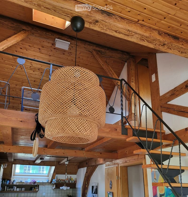 A modern, open-plan living space with exposed wooden beams and a large woven pendant light hanging from the ceiling. The area includes a staircase leading to an upper level, a kitchen with white tiles and wooden cabinets, and a small seating area with a f
