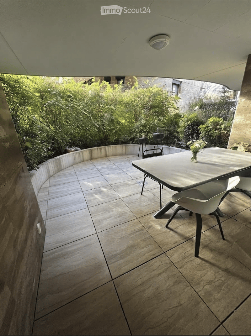 A modern, curved balcony with tiled flooring, a white table, chairs, and a flower vase, surrounded by lush greenery.