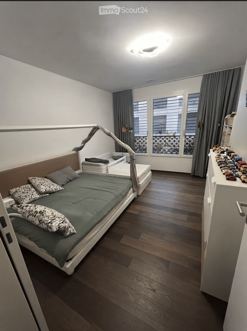 A modern bedroom with a large bed, patterned pillows, a nightstand, and a shelf with toys. The room features wooden flooring, large windows with curtains, and a cozy ambiance.