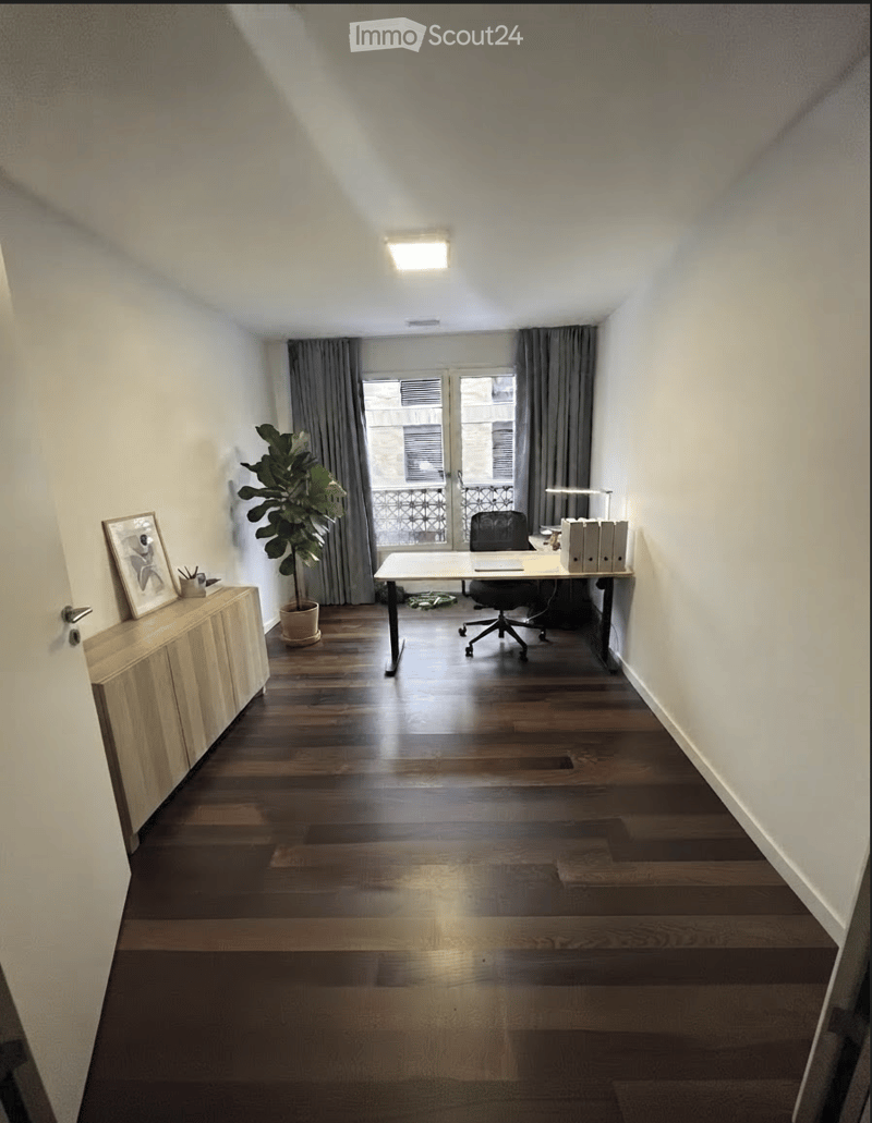 Modern office space with dark wooden flooring, white walls, a large window with grey curtains leading to a balcony, a desk with a chair, a cabinet with a picture frame, and a potted plant.