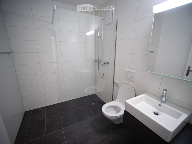 Modern bathroom with white tiles, a toilet, a sink, a shower, and a large mirror with built-in lighting.