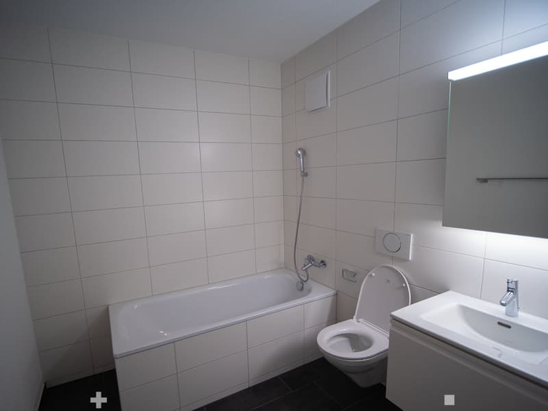 Modern bathroom with white tiled walls, a bathtub, toilet, sink, and a mirror with built-in lighting.