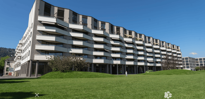Modern multi-story residential building with staggered balconies and large windows, surrounded by a green lawn.