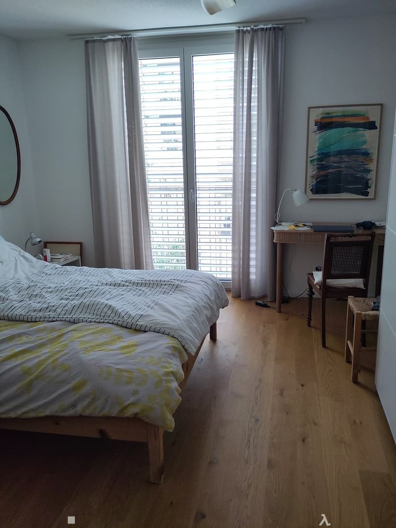 Cozy bedroom with wooden flooring, a double bed with striped and floral bedding, a desk with a chair, a mirror, and a lamp on the wall.