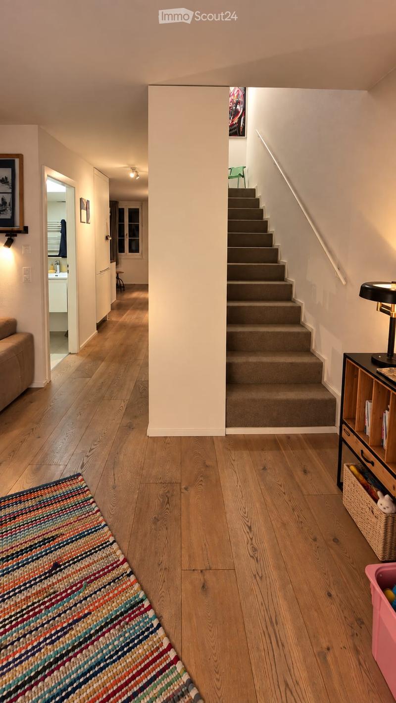 A well-lit hallway with wooden flooring, a staircase leading upwards, and a colorful runner rug. Visible rooms include a living area with a sofa and a kitchen area with cabinets and a sink.