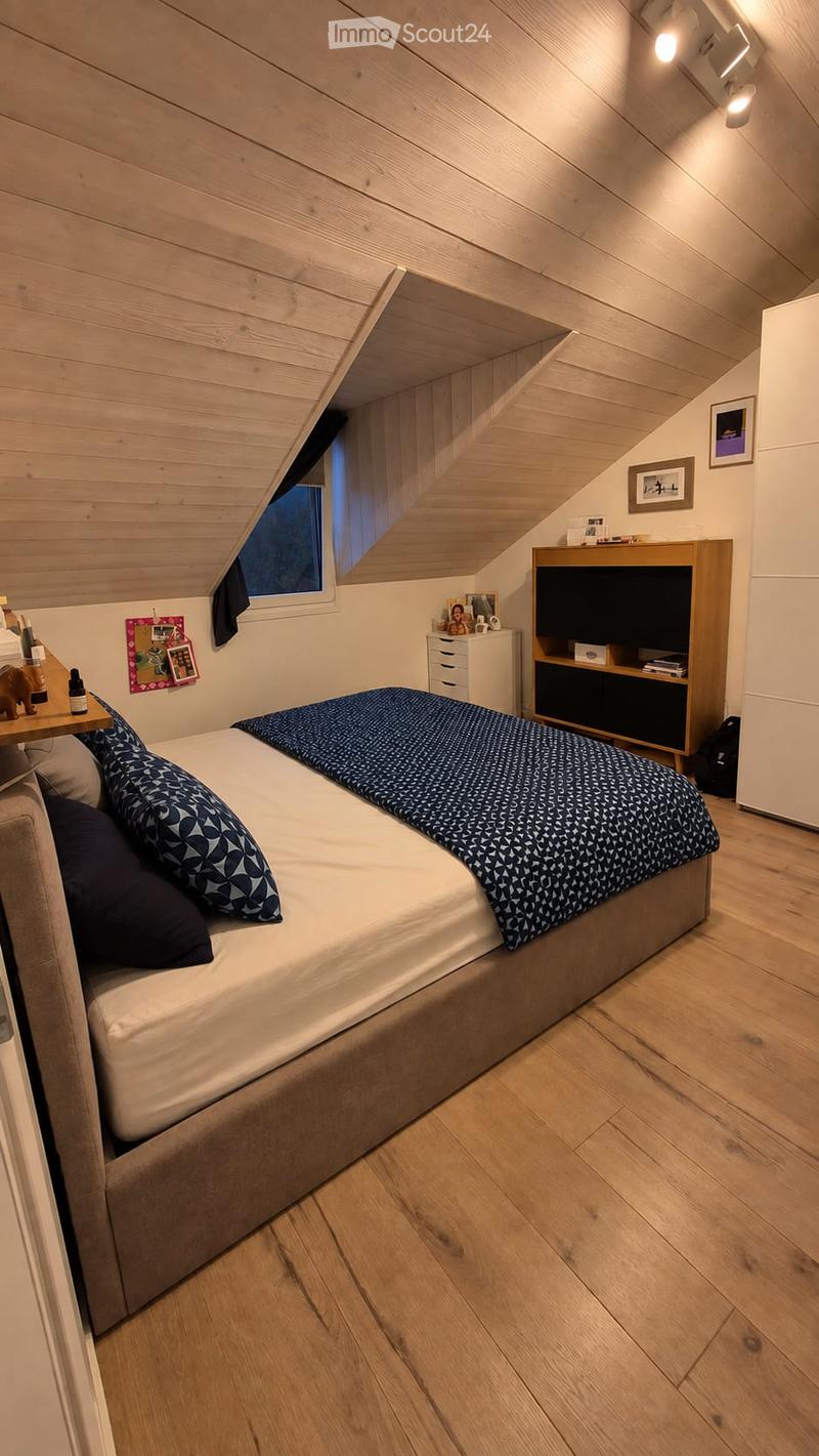 Cozy attic bedroom with wooden walls and ceiling, featuring a large bed with patterned bedding, a nightstand, a drawer unit, a bookshelf, and a small table with decorative items.