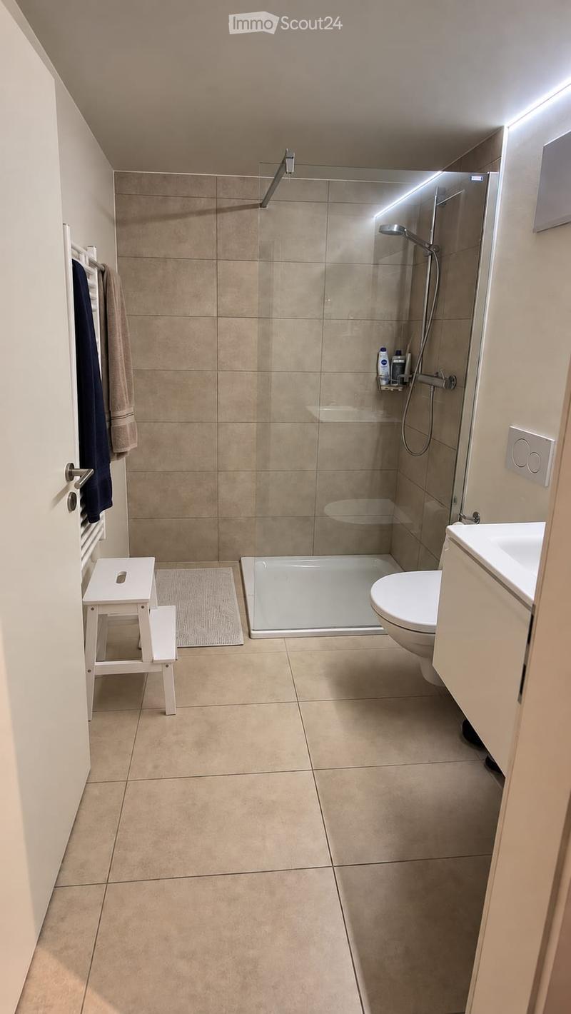 Modern bathroom with beige tiles, a glass-enclosed shower, a toilet, a small white stool, a towel rack with towels, and a shower shelf with toiletries.