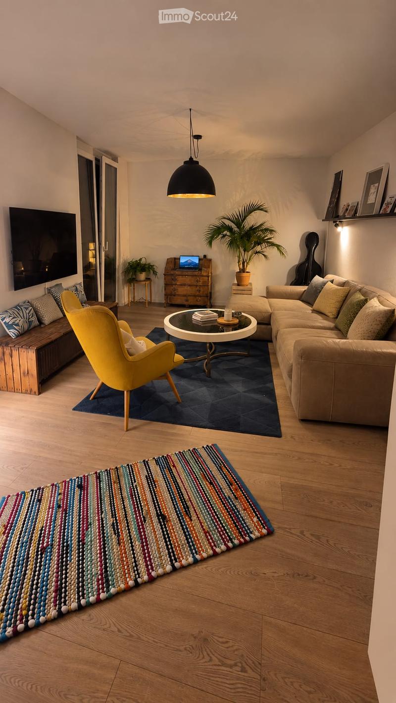 Modern living room with wooden flooring, a large sectional sofa, a yellow armchair, a round coffee table, a laptop on a small table, a potted plant, a guitar, and a colorful beaded runner rug.