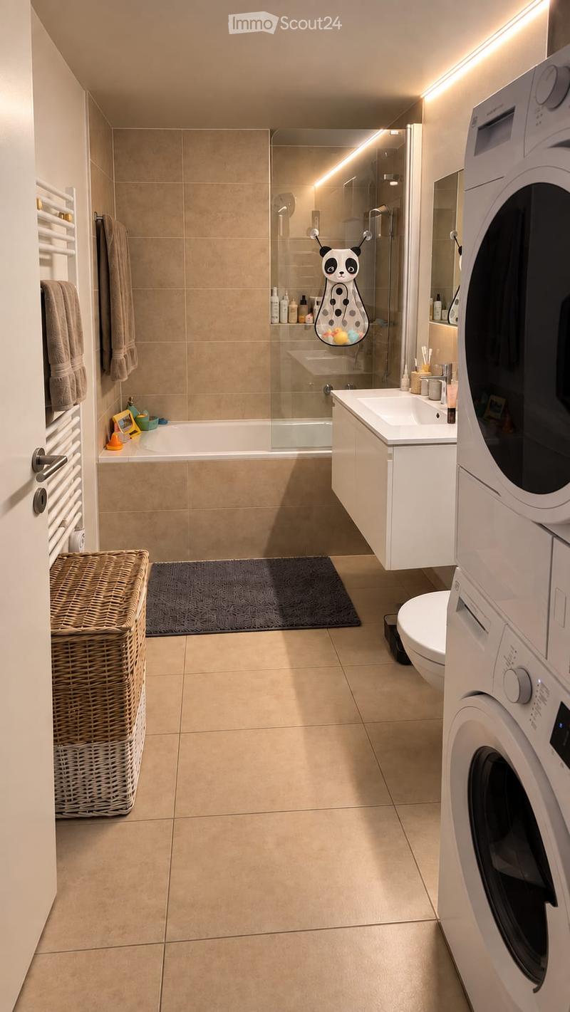 Modern bathroom with beige tiles, a bathtub, a sink, a toilet, a washing machine, a wicker basket, and a towel rack. The shower area features a glass enclosure with a decorative panda shower curtain.