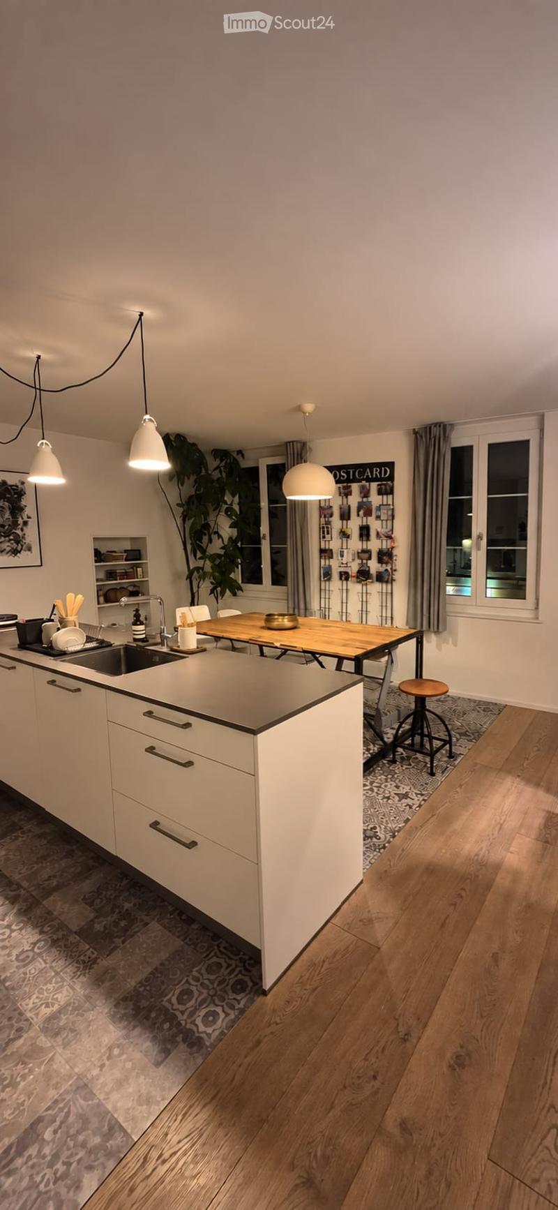 Modern kitchen with white cabinets, a central island, pendant lights, and a dining area with a wooden table and stool. Features a large window with a plant and a shelf with decorative items.