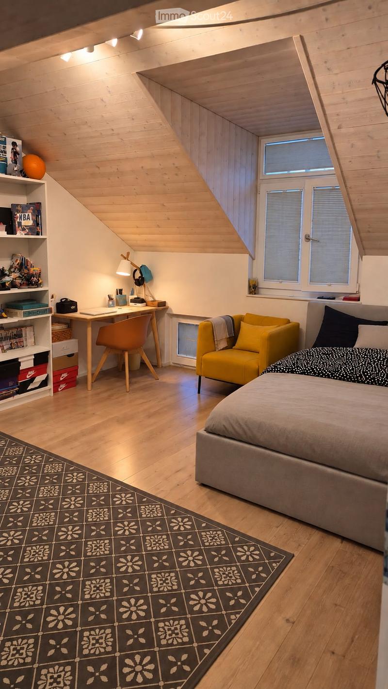 Cozy attic bedroom with wooden slanted ceilings, large windows, a bed, a yellow armchair, a desk with a chair, a shelf with books and decorative items, and a patterned rug.