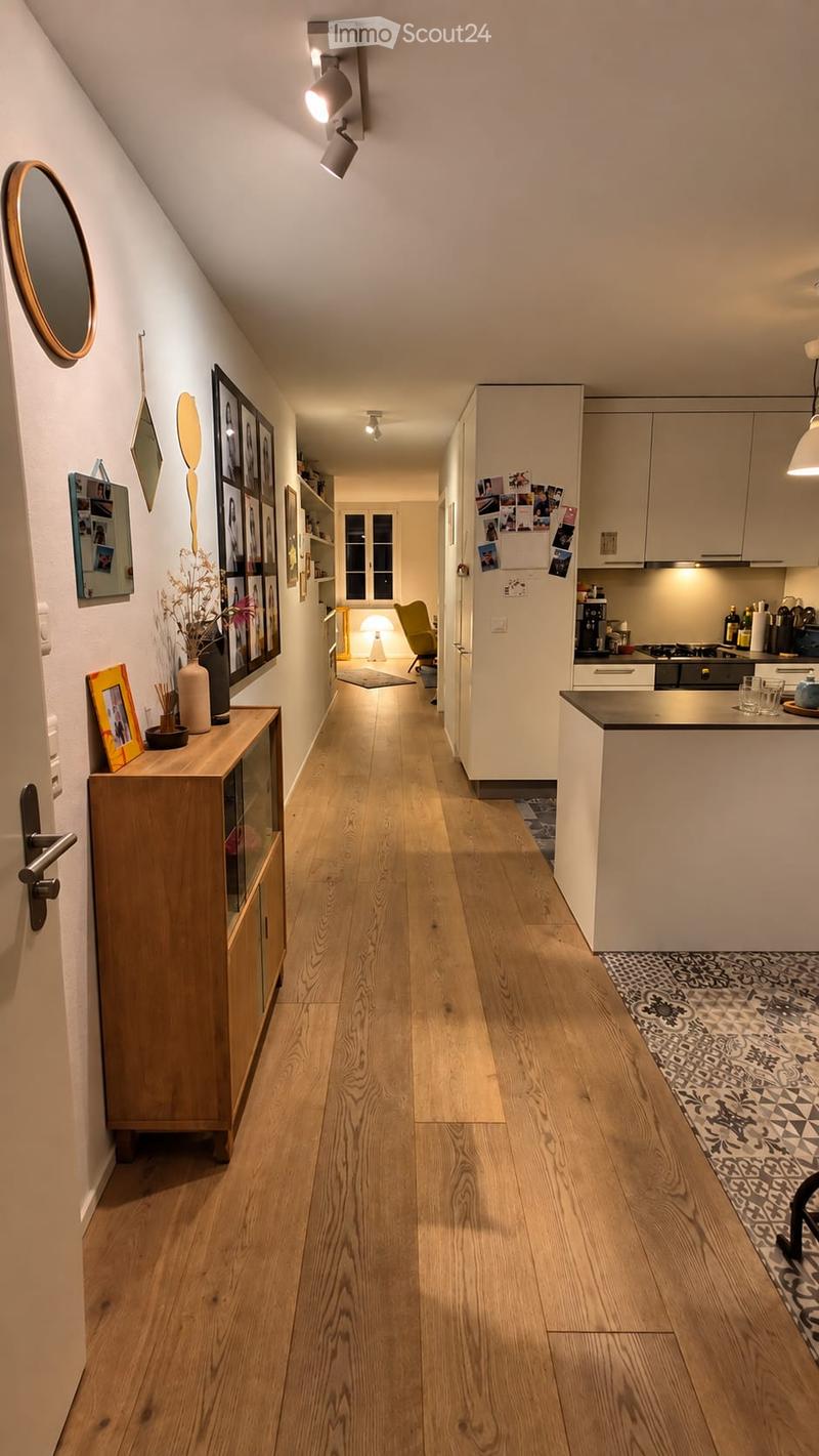 A well-lit hallway with wooden flooring, white walls, and modern decor including framed pictures, a small cabinet, and a vase with dried flowers. The hallway leads to a kitchen area with white cabinets and a black countertop.