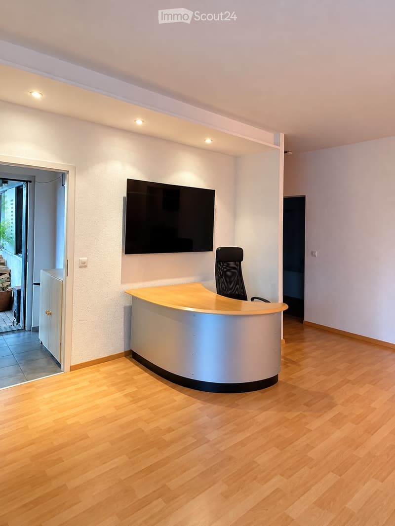Modern reception area with a curved reception desk, a black office chair, and a large wall-mounted screen. The room features light wooden flooring and recessed lighting.