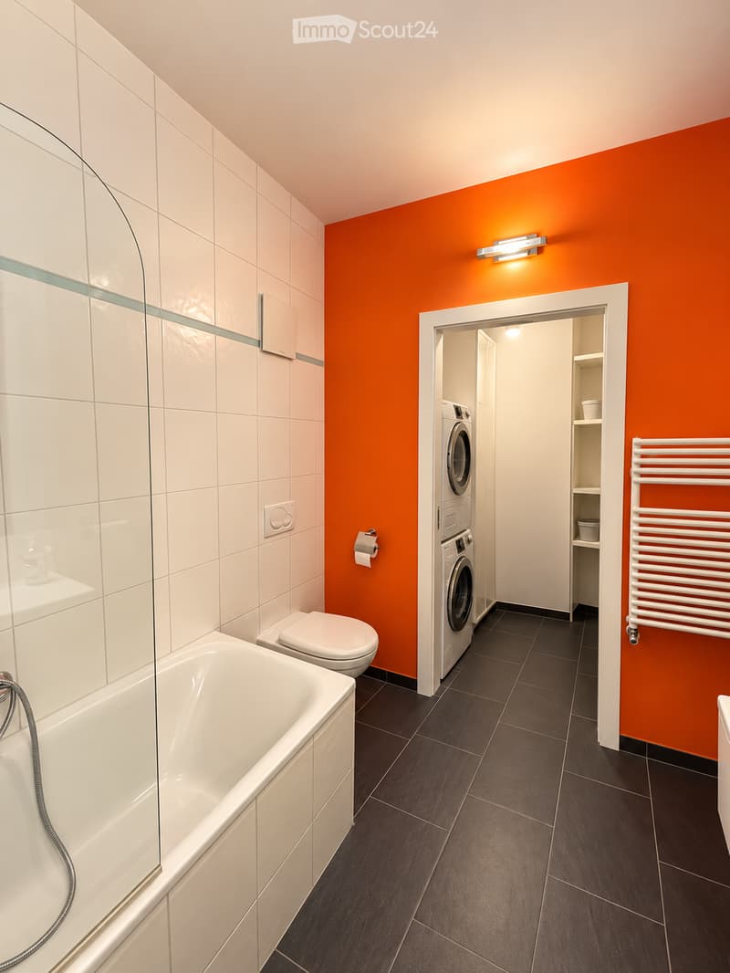 Modern bathroom with white tiles, dark floor, bathtub, toilet, washing machine, radiator, and shelf.