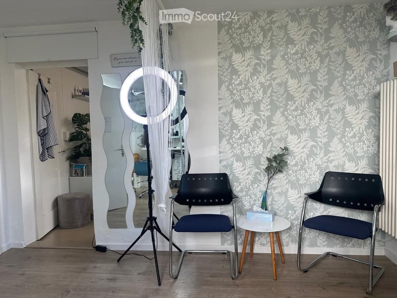 A stylish room with patterned wallpaper, two modern chairs, a small round table, a large mirror, a tripod, and a hanging plant. The room features a grey ottoman and a radiator.
