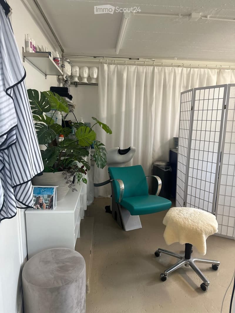 A well-lit salon or beauty room with a teal chair, a white stool, a large leafy plant, shelves with bottles and mannequin heads, and a curtained area.