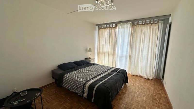 Cozy bedroom with wooden flooring, a double bed with dark bedding, a nightstand with a lamp, a small table with items, and a window with curtains.