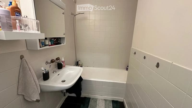 A small bathroom with a white tiled wall, a sink, a bathtub, a mirror, a cabinet, and a towel hanging on the wall.