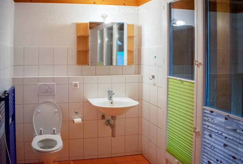 A compact bathroom with white tiled walls, a toilet, a sink with a chrome faucet, a mirror with wooden cabinets, and a glass shower enclosure with green blinds.