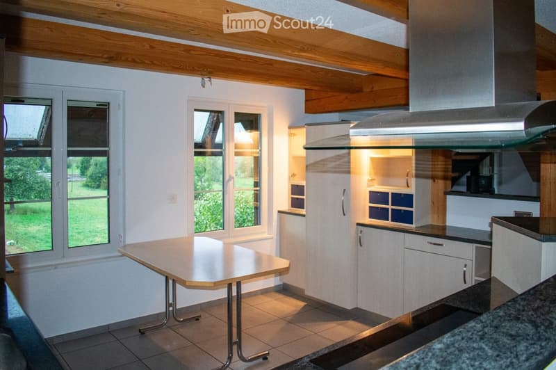 Bright kitchen with white cabinets, a central table, and large windows offering a view of the greenery outside. The room features a modern range hood and tiled flooring.