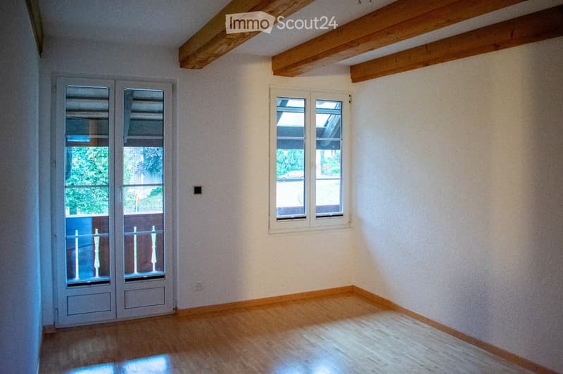 Empty room with wooden flooring, white walls, exposed wooden beams on the ceiling, and large windows with white frames. The room has a door with glass panels leading to a balcony area.