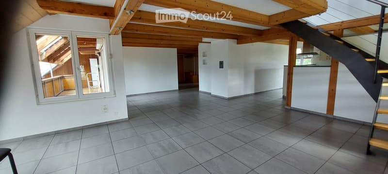 Empty room with exposed wooden beams, tiled floor, and large window showing a view of another room. No furniture or equipment visible.