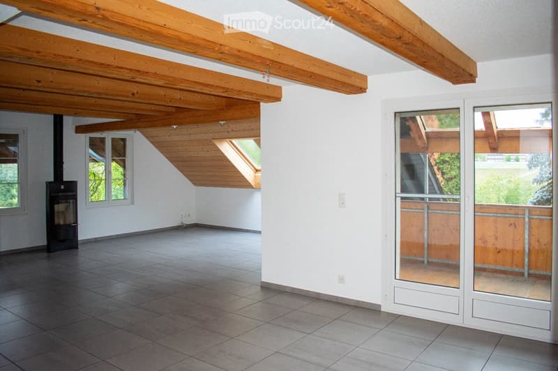 Empty room with exposed wooden beams, tiled floor, white walls, and large windows with a view of greenery. Features a fireplace and a sliding glass door leading to an outdoor area.