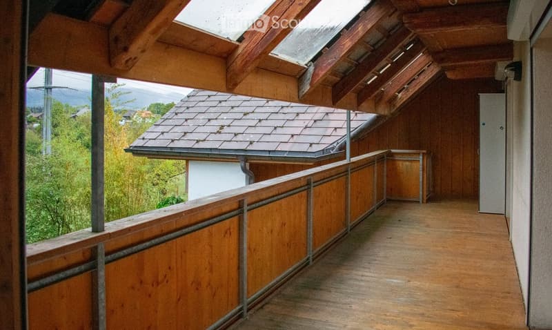 Wooden balcony with metal railing, skylight roof, and view of greenery and distant mountains.