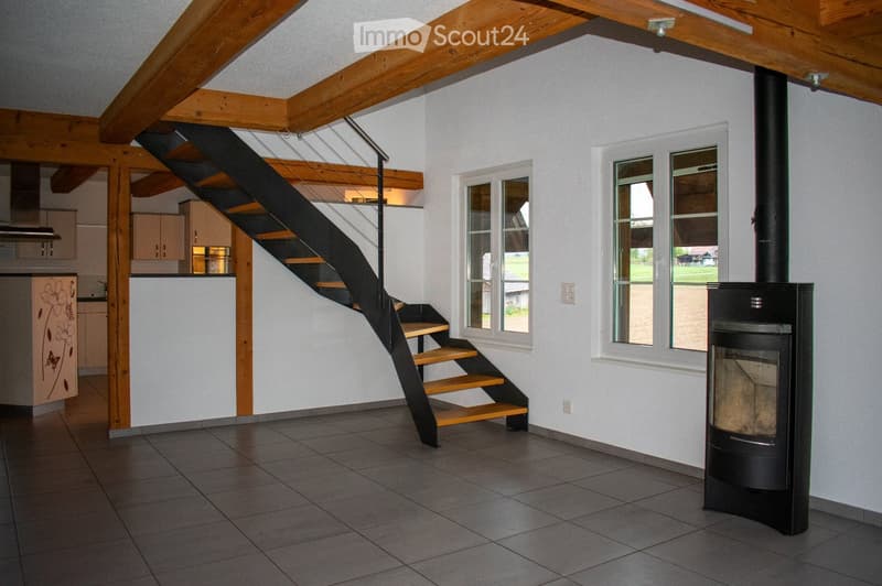 Modern interior with exposed wooden beams, a sleek black staircase, a fireplace, and a visible kitchen area with cabinets.