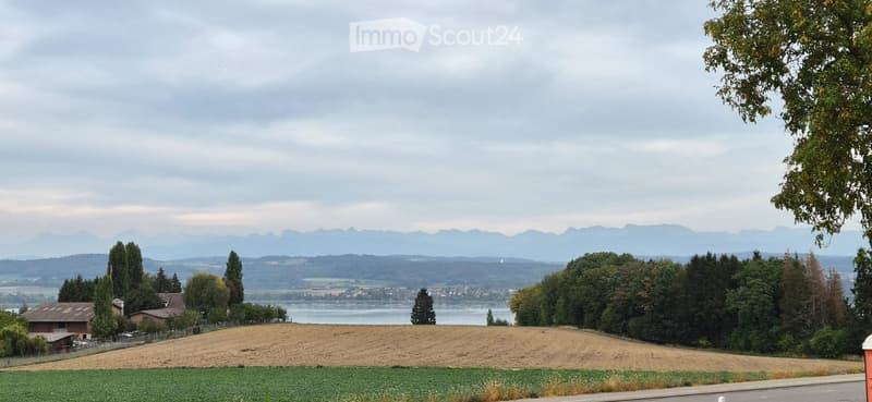 Scenic view of a rural landscape with fields, trees, a body of water, and distant mountains under a cloudy sky.