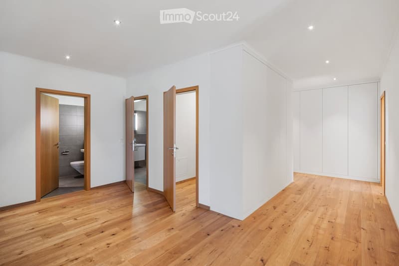Empty room with wooden flooring, white walls, and built-in white closet units. Two open wooden doors lead to adjacent spaces, including a bathroom with a toilet and sink.