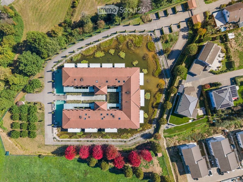 Aerial view of a large, modern house with a red-tiled roof, surrounded by a landscaped garden with a pond, pathways, and neighboring houses.