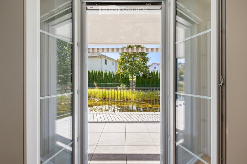View through a large glass sliding door onto a tiled balcony with a metal railing, overlooking a landscaped garden with water features and trees.