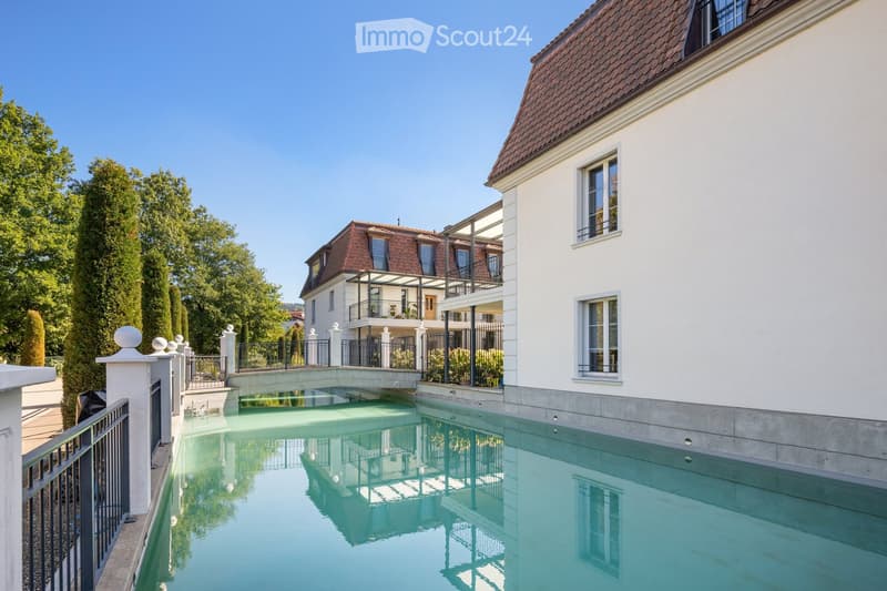 A modern outdoor area featuring a swimming pool with a clear blue water surface, surrounded by a white fence and lush greenery, with a contemporary house in the background.