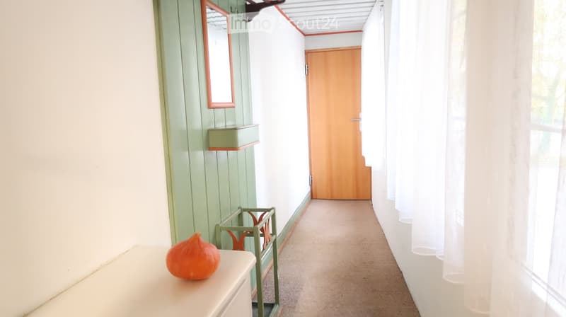A narrow hallway with light green and white walls, a wooden door at the end, a small shelf with a mirror, and a small table with an orange fruit on it.