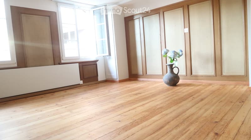 Empty room with wooden flooring, large windows, and a single decorative vase with flowers on the floor.