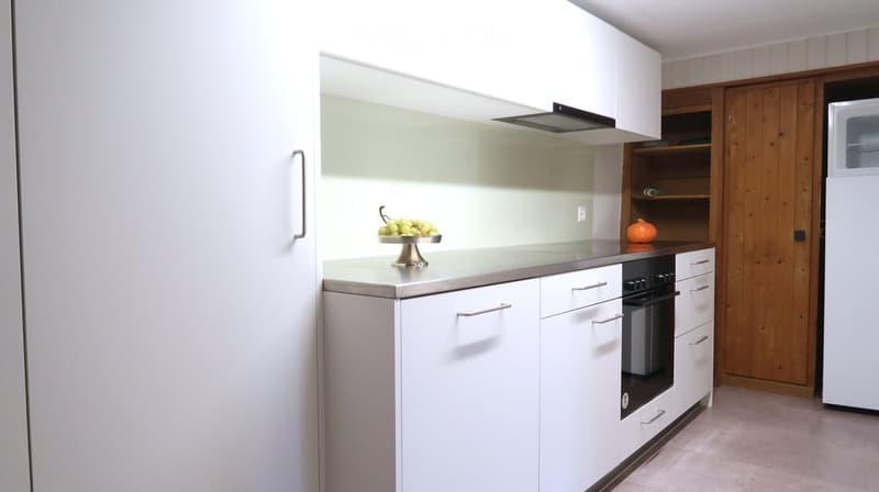 Modern kitchen with white cabinets, stainless steel countertops, built-in oven, and a small fruit bowl on the counter.